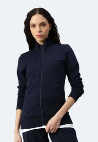 Giacca zip-up navy con colletto alto, dotata di due tasche laterali e polsini a coste. Realizzata in materiale morbido con un design aderente.