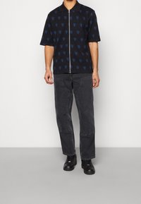 Camicia nera con zip a maniche corte, decorata con un motivo a cuori blu, abbinata a jeans neri larghi e sneakers nere.
