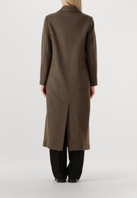 Lang brun frakke lavet af tekstureret stof, med en klippet krave og en midterste rygventil. Parret med sorte bukser og sko.