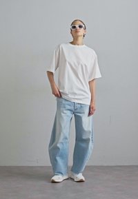 Tricou alb supradimensionat, blugi largi de culoare albastru deschis și adidași albi. Model purtând ochelari de soare albi; design minimalist, fără modele vizibile.