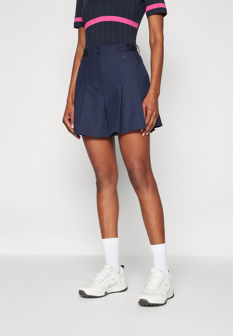 Falda corta plisada color navy hecha de tela ligera, con una cinturilla con cierre de botón y una textura suave. Combinada con zapatillas blancas.