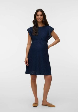 Uśmiechnięta kobieta w ciąży z długimi ciemnymi włosami, ubrana w granatową sukienkę z oczkami i jasnobrązowe wsuwane baleriny, stojąca na prostym tle.