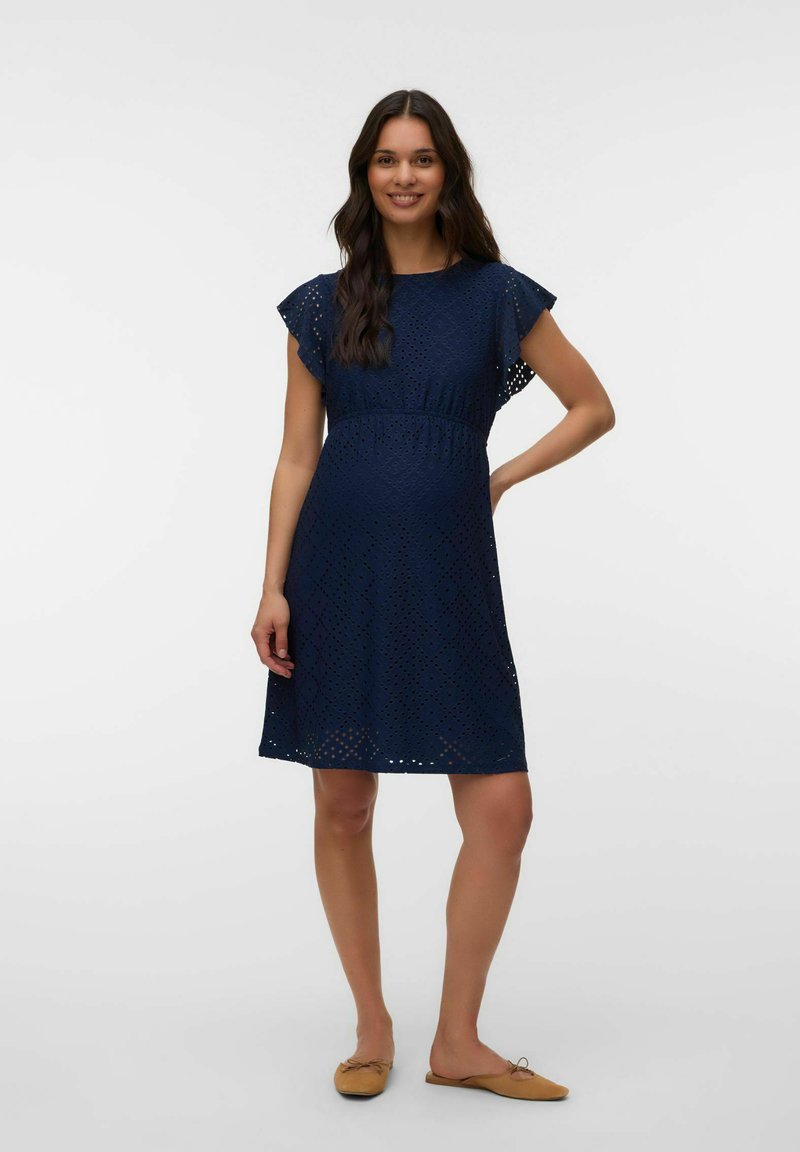 Femme enceinte souriante aux longs cheveux bruns foncés portant une robe bleu marine à œillets et des ballerines marron clair, debout devant un fond uni.