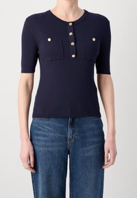 Granatowy prążkowany top z krótkim rękawem i okrągłym dekoltem, ozdobiony złotymi guzikami i dwiema kieszeniami na piersi. Faktura tkaniny jest gładka i dopasowana.