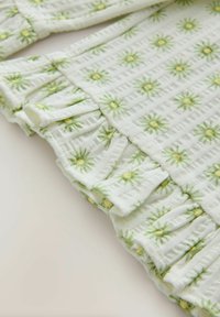 Tissu blanc texturé avec des bords froncés, présentant un motif floral répété en vert et jaune.