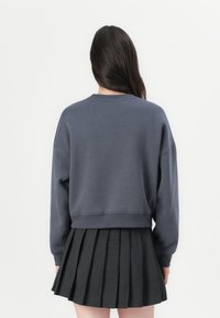 Sweatshirt cinza, curto, com mangas longas, apresentando um decote e punhos canelados, combinado com uma saia preta plissada.