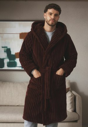 Mann trägt einen dunkelbraunen texturierten Bademantel mit den Händen in den Taschen und steht in einem modernen Wohnzimmer mit Sofa und abstrakter Wandkunst.