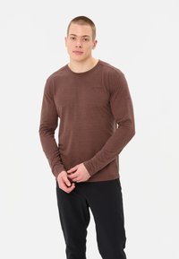 Lange mouwen, bruine t-shirt gemaakt van lichtgewicht stof, met een ronde halslijn en een logo detail op de borst. Gecombineerd met donkere broeken.