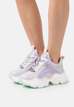 Gambe che indossano scarpe da ginnastica chunky in tonalità pastello viola e bianco, con suole bianche spesse e ciondoli a catena argentati sui lacci, su sfondo bianco.