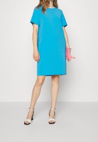 Modré šaty s krátkým rukávem a kulatým výstřihem, rovného střihu, doplněné o bílé a černé sandály na blokovém podpatku a růžovou kabelku.