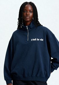 Sweat-shirt en polaire bleu marine avec un col haut, une demi-fermeture éclair et le texte blanc "c’est la vie" sur la poitrine. Coupe ample avec poignets et ourlet côtelés.