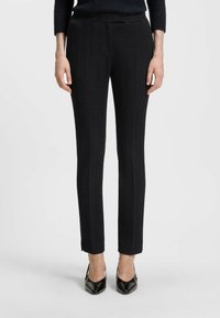 Mustat räätälöidyt housut, joissa on herringbone-kuitu, slim fit ja pieni halkio nilkassa, yhdistettynä teräväkärkisiin mustiin korkokenkiin.