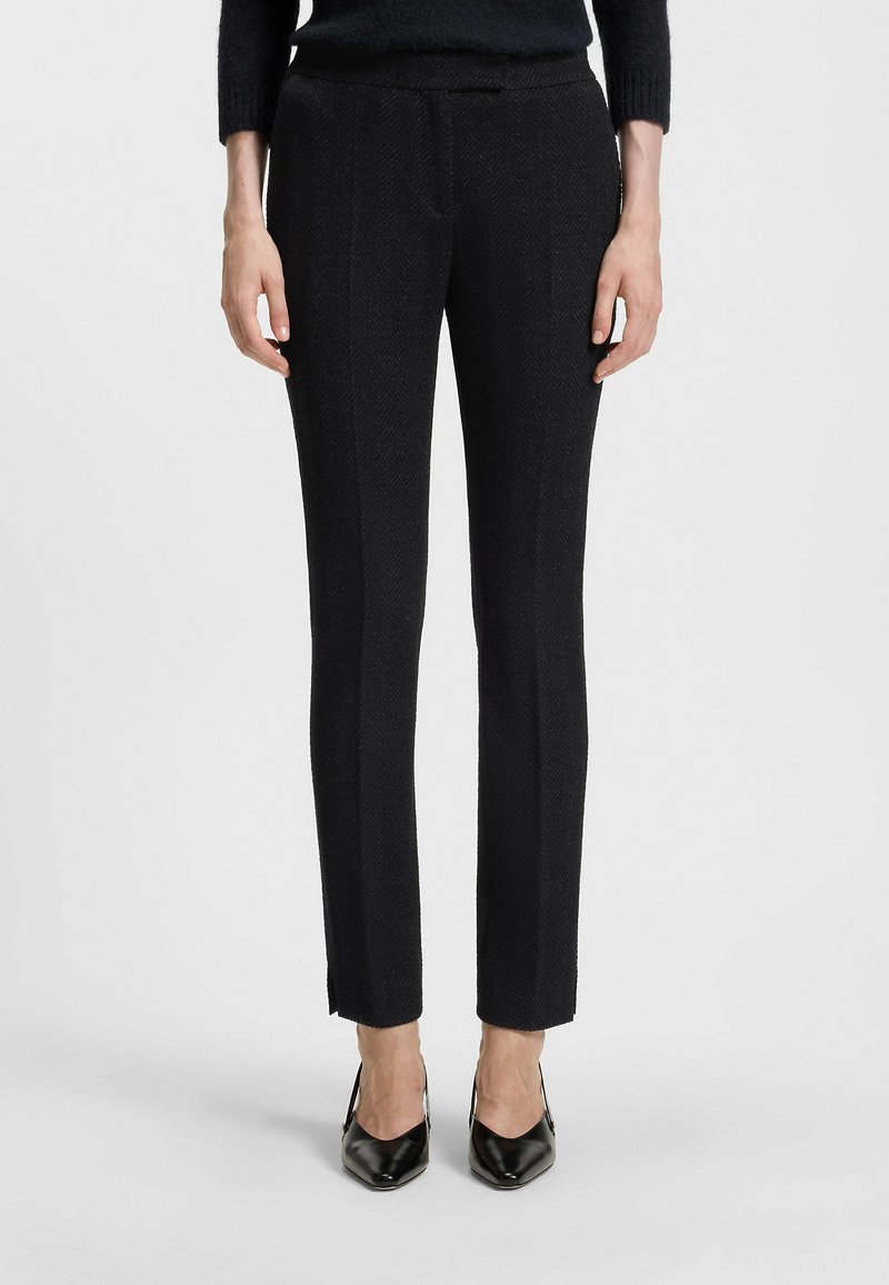 Mustat räätälöidyt housut, joissa on herringbone-kuitu, slim fit ja pieni halkio nilkassa, yhdistettynä teräväkärkisiin mustiin korkokenkiin.