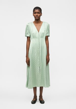 Nő világoszöld, texturált midi ruhát visel, amelyen pöttyös rövid ujj van, mély V-nyakkivágás, elöl anyaggal borított gombok és fekete balerinacipők.