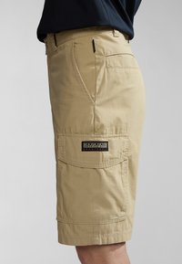 Pantaloni cargo beige realizzati in misto cotone, con tasche laterali, passanti per cintura e una patch logo cucita sulla coscia sinistra. Texture liscia, lunghezza al ginocchio.