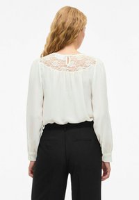 Weiße Bluse mit Spitzenverzierung am Rücken, langen Ärmeln und Schlüssellochdetail. Kombiniert mit schwarzen, taillierten Hosen. Glatte Stoffoberfläche.