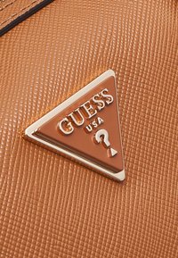 Brązowa skórzana powierzchnia o fakturze z trójkątnym metalowym logo Guess USA z uniesionym symbolem znaku zapytania.