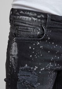 Jeans negre din denim cu un design deteriorat, cu bucăți destrămate și un model de stropi în nuanțe de gri, evidențiind textura și detaliile.