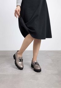 Mocasines gris metálico con suelas negras gruesas y detalles de cadena, combinados con una falda negra hasta la rodilla. Textura suave y diseño moderno.