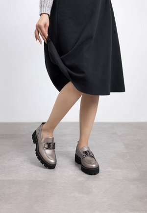 Mocasines gris metálico con suelas negras gruesas y detalles de cadena, combinados con una falda negra hasta la rodilla. Textura suave y diseño moderno.