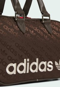 Brown Adidas bag with repeated logo pattern and large white embroidered Adidas text, featuring dark leather handles and silver metal hardware.