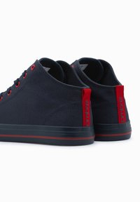 Marineblaue Canvas-Sneaker mit roten Akzenten und Ösen, ausgestattet mit einer flachen schwarzen Sohle und einem roten Fersenabsatz mit dem Markennamen.
