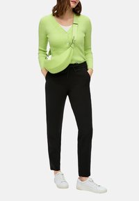 Cardigan verde a coste con bottoni, pantaloni neri e sneakers bianche. Piccola borsa a tracolla verde. Outfit semplice e casual con accenti vivaci.
