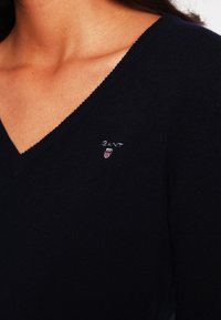 Granatowy sweter w serek wykonany z miękkiego materiału, z małym haftowanym logo na piersi. Gładka tekstura i dopasowany krój.