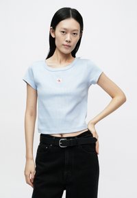 Light blue ribbed t-shirt with short sleeves, round neckline, and a small red logo patch on the chest, paired with black high-waisted jeans.