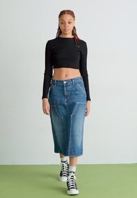 Denim midi sukňa v modrej farbe s charakteristickým šitím, prednými vreckami a rozparkom, skombinovaná s čiernym rebrovaným crop topom a vysokými teniskami.