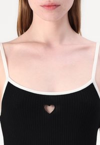 Svart ribbet tanktop med hvit kant, med et hjerteformet utsnitt på brystet. Stroppene er tynne, og teksturen er glatt.