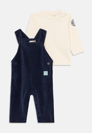 Salopette in corduroy blu navy con bretelle regolabili e due tasche laterali, abbinata a una maglietta a maniche lunghe color crema con scollatura rotonda.