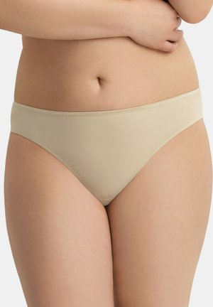 Femme portant un sous-vêtement style bikini beige sans coutures, un bras posé sur la taille.