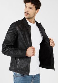 Zwarte leren jas met gequilte schouderdetails, ritssluiting en zijzakken. Binnenvoering is zwart. Gedragen over een wit shirt.