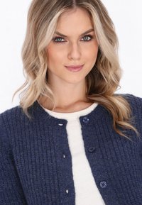 Granatowy sweter zapinany na guziki, z teksturowanym wzorem, okrągłym dekoltem i niebieskimi guzikami. Nosi się go na białej bluzce, która ma dopasowany krój.
