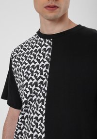 Schwarzes T-Shirt mit einem geometrischen weißen Muster auf einer Seite. Rundhalsausschnitt, kurze Ärmel und aus weichem Baumwollmaterial.