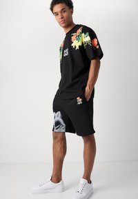 T-shirt en coton noir avec des impressions florales colorées sur les manches, logo sur la poitrine ; associé à un short noir avec un design graphique gris. Baskets blanches.