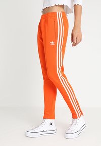 Henkilö, joka on pukeutunut kirkkaan oransseihin Adidas-vyölenkkeihin, joissa on valkoiset raidat, ja valkoisiin korkeisiin Converse-tennareihin, seisoo yksinkertaista taustaa vasten.