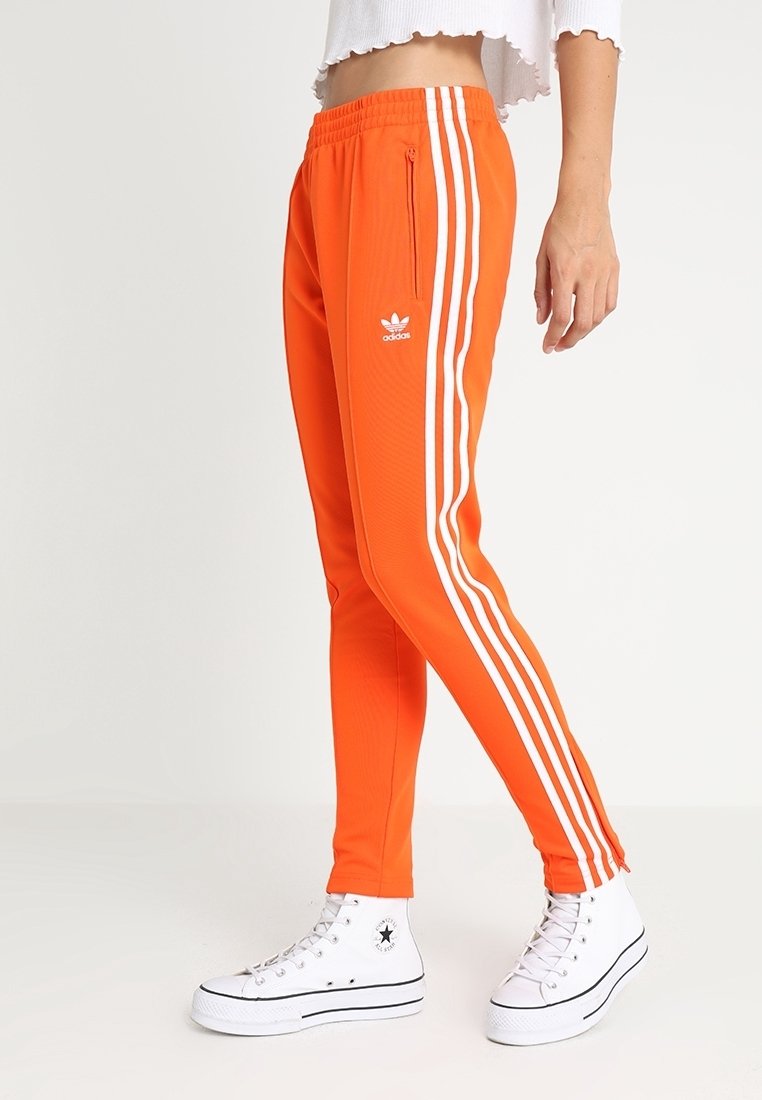 Henkilö, joka on pukeutunut kirkkaan oransseihin Adidas-vyölenkkeihin, joissa on valkoiset raidat, ja valkoisiin korkeisiin Converse-tennareihin, seisoo yksinkertaista taustaa vasten.