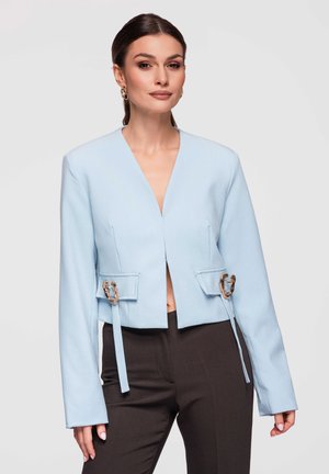 Kvinde iført en lyseblå cropped blazer med guldlås-detaljer og sorte bukser, poserer mod en ensfarvet hvid baggrund.