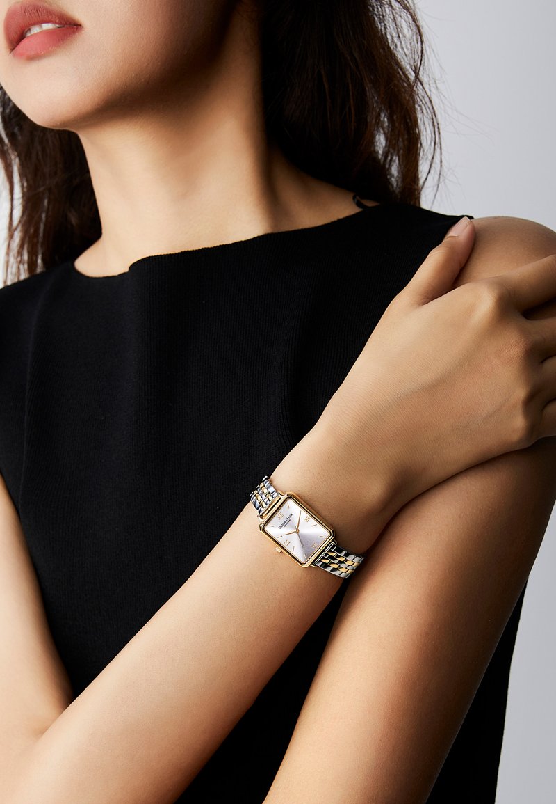 Žena v černém tílku, s překříženýma rukama, ukazující dvoubarevné kovové náramkové hodinky se čtvercovým zlatým rámečkem a bílým ciferníkem.