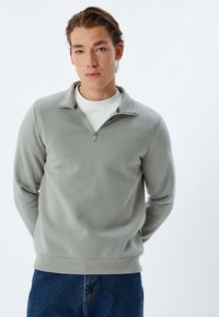 Felpa grigia chiara con zip a quarto realizzata in tessuto morbido, caratterizzata da orlo a coste e maniche lunghe, abbinata a jeans blu.