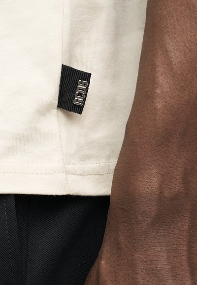 Κοντινό πλάνο στο τελείωμα ενός μπεζ πουκαμίσου με μια μαύρη ετικέτα υφάσματος που γράφει "GCDS" δίπλα στο χέρι ενός ατόμου με σκουρόχρωμο δέρμα και μαύρο παντελόνι.