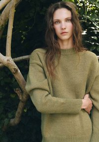 Maglione lavorato a maglia verde oliva con un collo alto, maniche larghe e polsini a costine, caratterizzato da un motivo testurizzato. Lo sfondo include una vegetazione naturale.