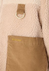 Veste en polaire beige doux avec une surface texturée, dotée d'une poche en velours côtelé marron et d'un accent en métal en forme d'anneau. Logo "Barbour" en doré.