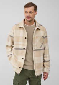 Man in een beige geruite knoopjass over een tan kleurige trui en olijfgroene cargobroek, staand tegen een effen lichtgrijze achtergrond.