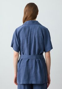 Frau mit langen braunen Haaren, von hinten gezeigt, trägt ein locker sitzendes, blaues Kurzarmhemd mit einem passenden Gürtel und blauen Hosen.