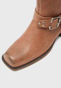 Bottine en cuir marron à bout carré avec une bride décorative comprenant une boucle métallique et des rivets sur fond blanc.