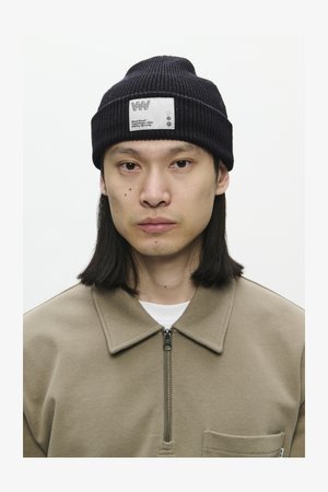 Ung person med glat skulderlangt hår iført en sort strikket hue med et hvidt mærke og en beige zip-up skjorte.