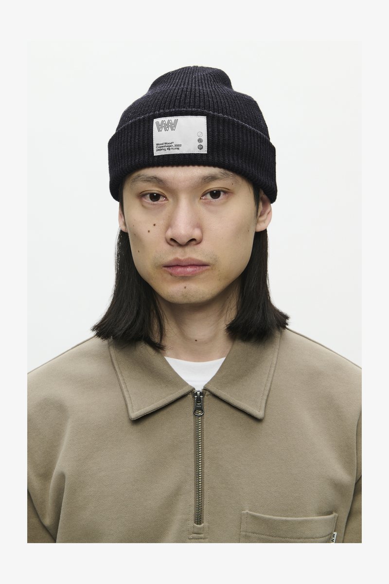 Fiatal személy, aki egyenes, vállig érő hajú, fekete kötött sapkát visel, rajta fehér címkével, és bézs színű, cipzáros pólót hord.